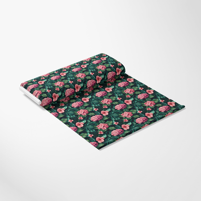A roll of fabric with a floral pattern featuring pink flowers and green leaves on a dark background, unrolled on a light gray surface.