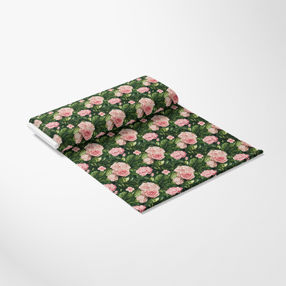 A roll of decorative fabric with a floral pattern of pink roses and green leaves on a dark background, partially unfurled on a white surface.